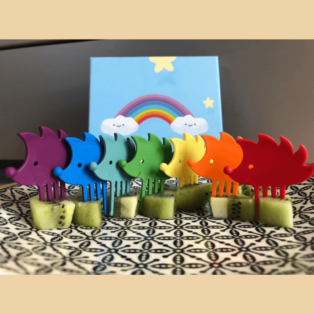 Set aus sieben Snack-Pieksern in Igel-Form in, jeder in einer Farbe des Regenbogens, in einer Reihe jeweils in ein Stück Kiwi gesteckt.