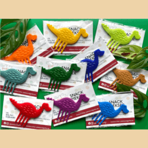 Blick von oben auf ein Set aus zehn Snack Pieksern in Dinosaurier-Form, jeder in einer anderen Farbe, einzeln verpackt und liegend vor grünem Hintergrund, dekoriert mit Pflanzen.
