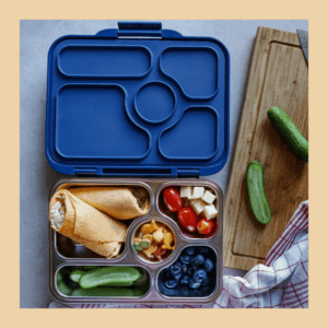 Zu sehen ist die Vesperdose "Yumbox Presto" aus Edelstahl mit fünf Fächern und einem dunkelblauem Deckel aus Silikon, gefüllt mit Wraps, Gurken, Blaubeeren, Tomaten, Käse und Knabbereien.