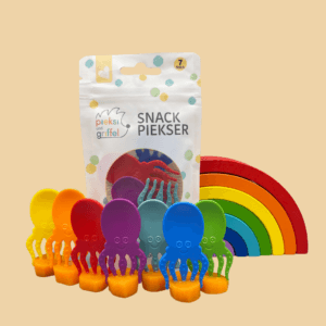 Zu sehen ist das "Ocean's Seven" Set der Snack Piekser Tintenfische von Pieksi und Griffel in Regenbogenfarben. Alle sieben stecken in einzelnen kleinen Karottenstücken und im Hintergrund ist die Verpackung sowie ein hölzerner, bunter Regenebogen zu sehen.